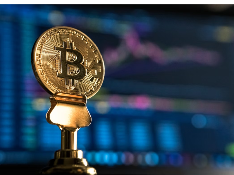 Na imagem enviada, vemos uma representação física do Bitcoin (em forma de uma moeda dourada com o símbolo ₿) em destaque, com telas de gráficos financeiros ao fundo. Essa cena remete diretamente ao universo das criptomoedas e ativos digitais.