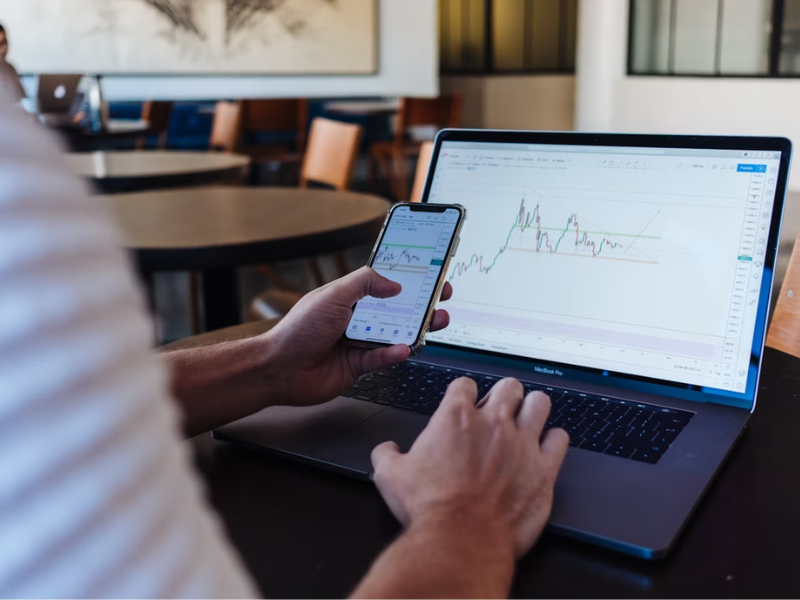 Na imagem vemos uma pessoa analisando gráficos financeiros em um laptop (MacBook Pro) e um smartphone. Ambos os dispositivos mostram gráficos de análise técnica, que são comumente utilizados para negociar ativos financeiros.