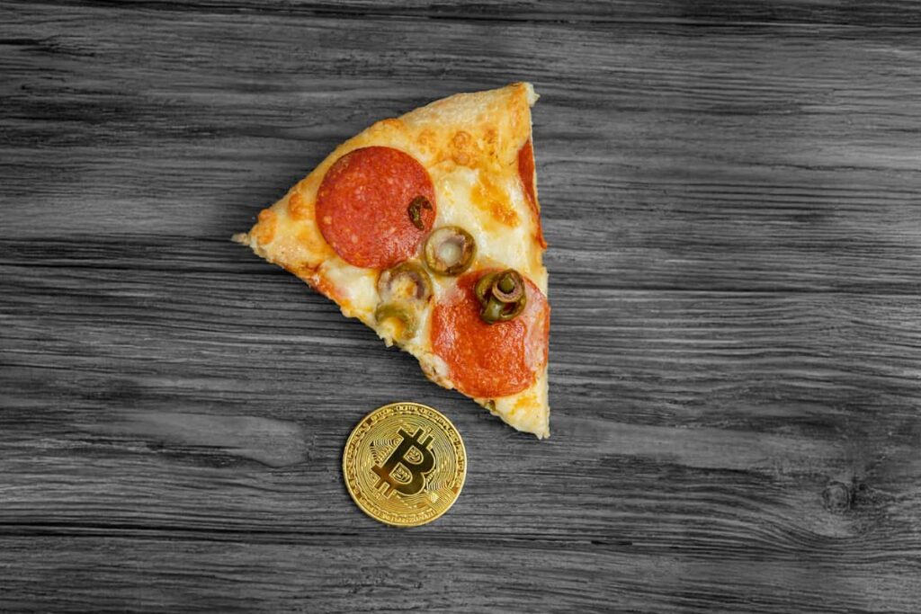 Fatia de pizza com pepperoni e azeitonas ao lado de uma moeda de Bitcoin sobre fundo de madeira, representando o Pizza Day.
