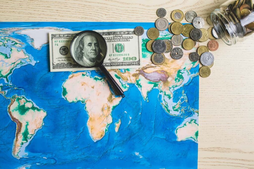 Mapa com cédula de dólar, lupa e moedas estrangeiras ilustrando o tema como investir no exterior morando no Brasil.