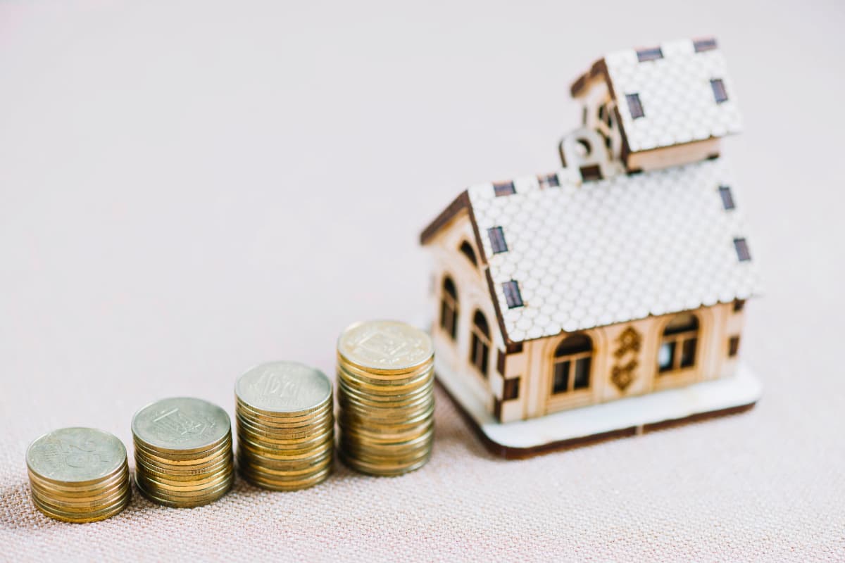 Imagem de uma casa pequena com pilhas de moedas em uma mesa, simbolizando investimentos em tijolo, papel e híbrido.