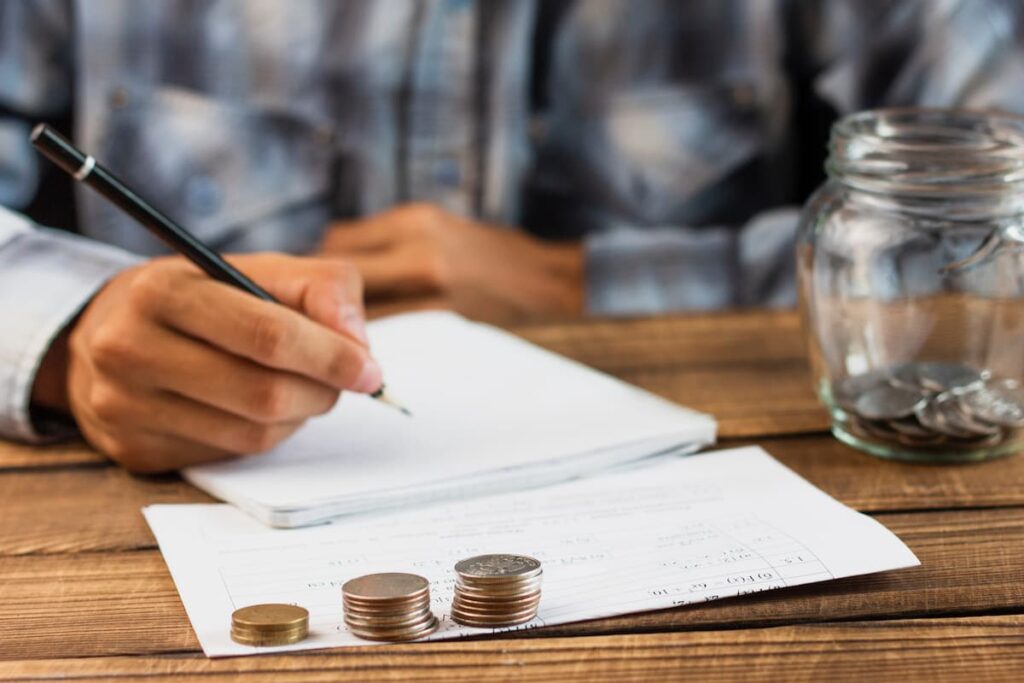 Pessoa anotando em caderno ao lado de moedas e jarro de troco, representando conceitos do glossário de investimentos.