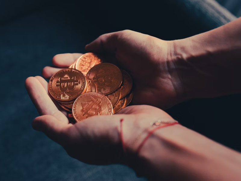 A imagem apresenta duas mãos humanas em posição de concha, segurando uma quantidade visível de moedas físicas douradas com o símbolo do Bitcoin (₿) em relevo.