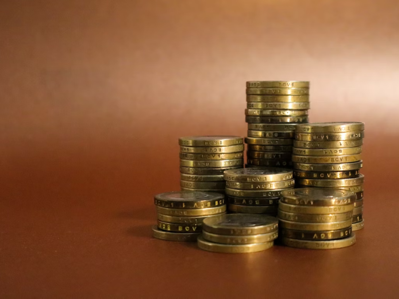 A imagem mostra várias pilhas de moedas organizadas em diferentes alturas, com um fundo marrom simples e neutro, que destaca o foco nas moedas. A composição transmite uma sensação de crescimento, estabilidade financeira e acúmulo de capital.