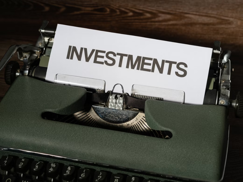 A imagem mostra uma máquina de escrever com uma folha de papel onde está escrita a palavra "INVESTMENTS" (investimentos). A composição sugere o tema de finanças e investimentos, utilizando um elemento vintage para destacar a importância atemporal do assunto.