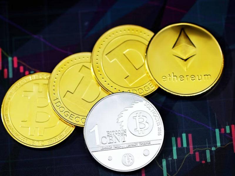 A imagem mostra cinco moedas físicas representando diferentes criptomoedas, colocadas sobre um fundo que exibe gráficos de velas (candlesticks) típicos de plataformas de negociação financeira.