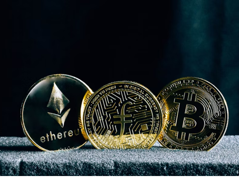 A imagem mostra três moedas físicas douradas que representam criptomoedas populares, dispostas lado a lado sobre uma superfície aveludada escura, com fundo preto fosco, criando um visual elegante e profissional.