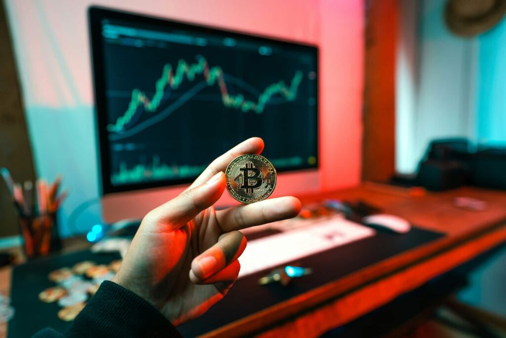 Pessoa segura uma moeda de bitcoin com gráfico no monitor ao fundo, representando investimentos em criptomoedas ou ações.