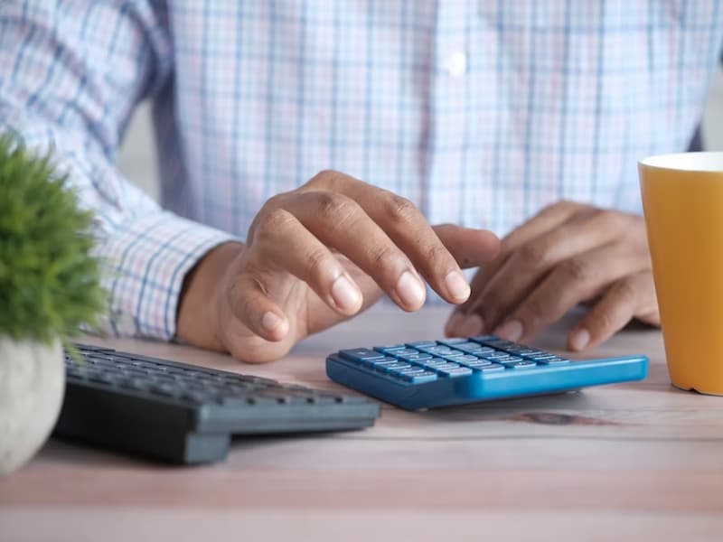 A imagem mostra as mãos de uma pessoa digitando em uma calculadora azul, sobre uma mesa de madeira clara. A pessoa veste uma camisa social xadrez, sugerindo um contexto profissional ou de negócios.