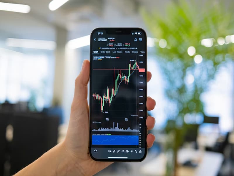 A imagem mostra uma mão segurando um smartphone que exibe uma plataforma de negociação de ativos financeiros, como ações ou criptomoedas. A tela apresenta um gráfico de velas japonesas (candlestick), muito comum em análises técnicas do mercado financeiro.