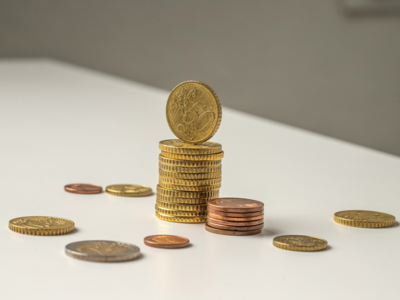A imagem mostra várias pilhas e moedas soltas dispostas sobre uma superfície branca. No centro, destaca-se uma pilha de moedas douradas (de 50 centavos de euro) com uma moeda posicionada verticalmente no topo, sugerindo destaque e crescimento. Ao lado, há uma pilha menor de moedas de cobre e outras moedas espalhadas, indicando diversidade de valores.