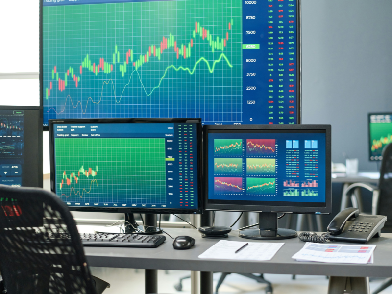 A imagem mostra uma sala de operações financeiras (também conhecida como trading room), com múltiplos monitores exibindo gráficos de preços e indicadores de mercado. Esse ambiente é típico de onde se negociam ativos financeiros em tempo real.