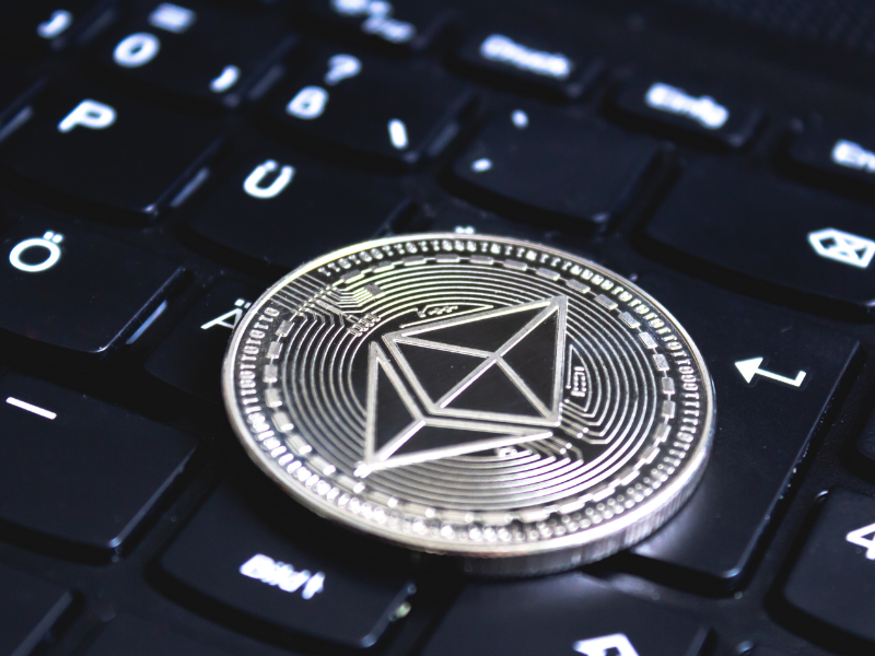 A imagem é um close-up focado em uma moeda digital (criptomoeda) de cor prateada, que está colocada sobre as teclas pretas de um teclado de computador ou laptop.