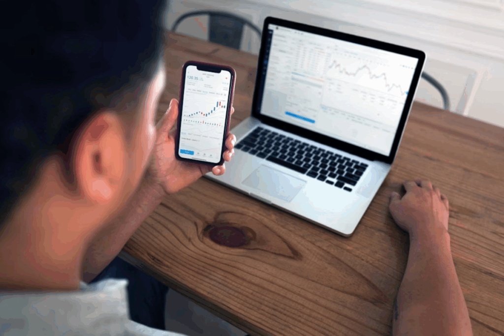 A imagem mostra uma pessoa sentada em uma mesa de madeira, focada em duas telas que exibem gráficos financeiros. Na mão esquerda, ela segura um smartphone com a tela ligada em um aplicativo de investimento, que mostra um gráfico de linha verde, indicando uma tendência de alta.