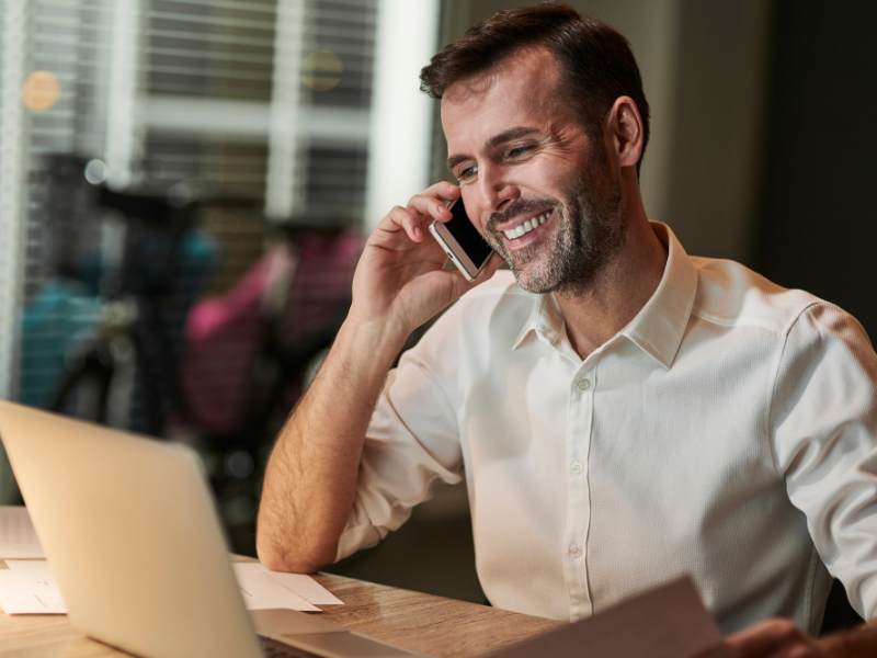 A imagem mostra um homem de meia-idade com barba, vestindo uma camisa social branca, sentado em uma mesa e conversando ao telefone. Ele está sorrindo, sugerindo uma conversa agradável. 