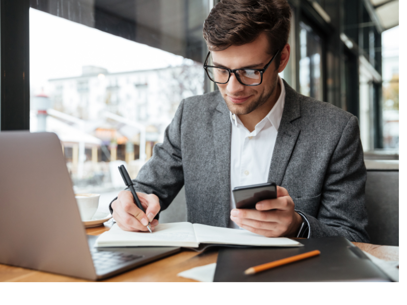 A imagem mostra um homem jovem de negócios trabalhando em um ambiente moderno, possivelmente um café ou coworking. Ele está anotando em um caderno, enquanto segura um celular e tem um laptop aberto à sua frente. O cenário transmite foco, produtividade e organização, típico de alguém lidando com planejamento financeiro, investimentos ou gestão patrimonial.