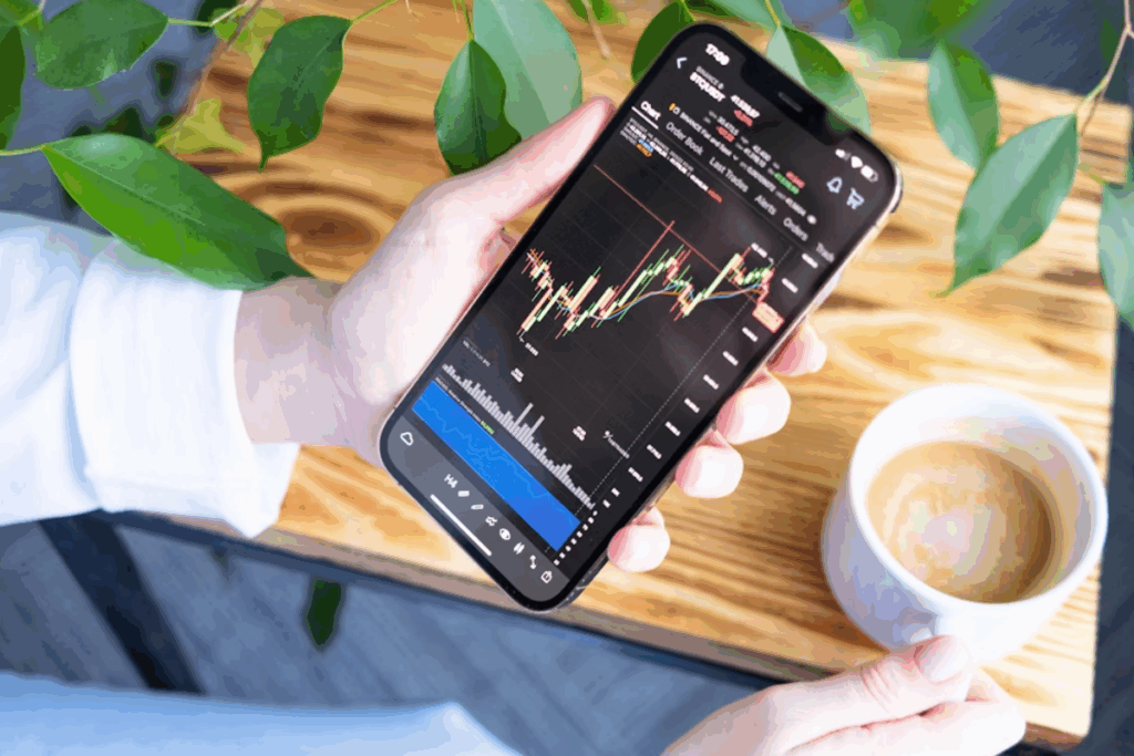 A imagem mostra a mão de uma pessoa segurando um smartphone sobre uma mesa de madeira. A tela do celular exibe um gráfico de mercado de ações ou criptomoedas.