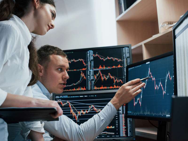 A imagem mostra um homem e uma mulher em um escritório, examinando múltiplos monitores de computador que exibem gráficos financeiros.