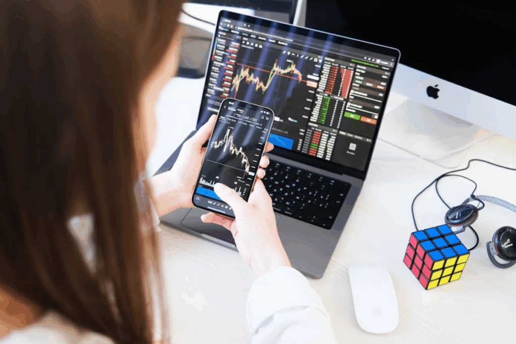 Na mesa, ao lado do notebook, há um cubo mágico, um mouse branco e um par de fones de ouvido com fio. Ao fundo, no lado direito, é possível ver um monitor de computador. A imagem sugere uma pessoa acompanhando o mercado financeiro, possivelmente de criptomoedas, em seus dispositivos.