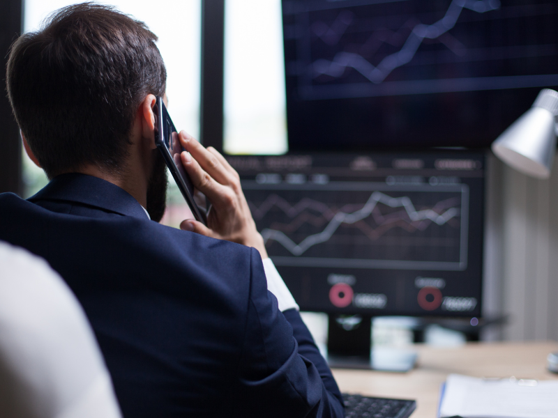 A imagem é uma vista traseira de um jovem empresário ou analista em um escritório, concentrado em uma chamada telefônica enquanto observa gráficos financeiros em um monitor de computador.