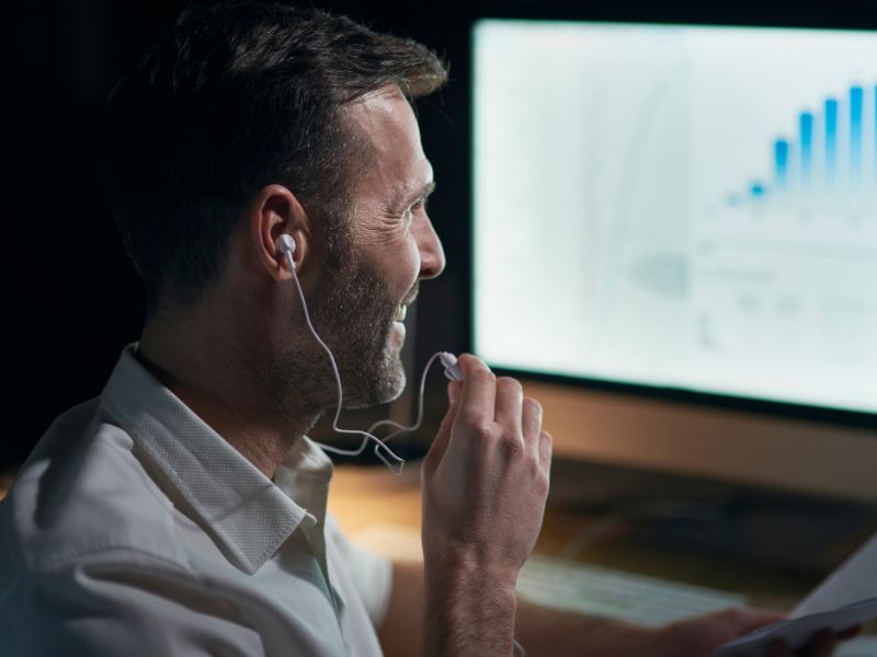 A imagem mostra um homem sorridente usando fones de ouvido enquanto trabalha em frente a um computador. Na tela, há um gráfico de barras ascendentes, indicando análise de dados ou investimentos.