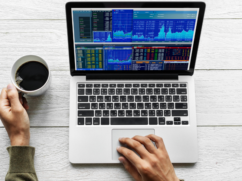 A imagem é uma vista superior que mostra uma pessoa interagindo com um laptop sobre uma superfície de madeira clara ou branca com textura de tábuas. A cena sugere um ambiente de trabalho focado em análise de dados ou mercado financeiro.