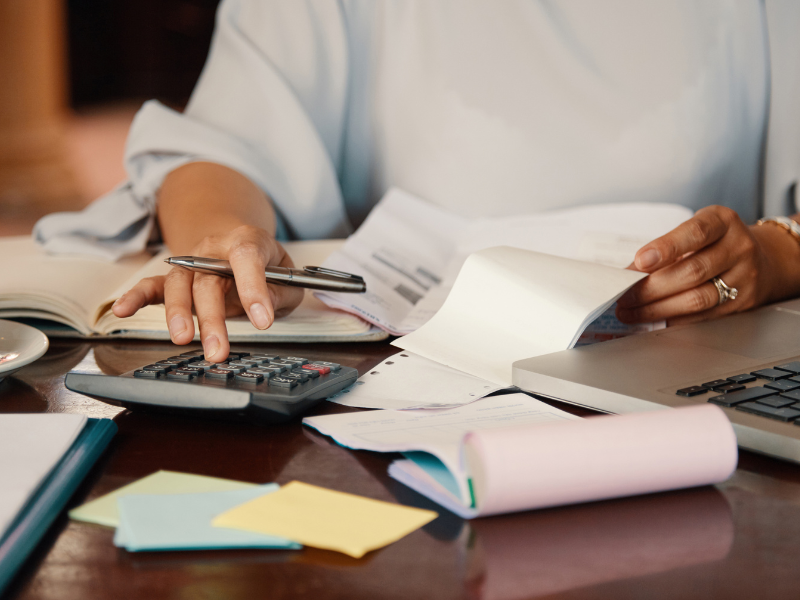 A imagem mostra uma pessoa trabalhando em uma mesa com laptop, papéis, calculadora e caneta, sugerindo uma atividade de contabilidade ou gestão financeira.