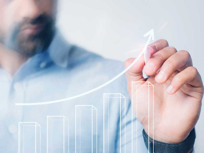 A imagem mostra um homem de camisa azul clara desenhando um gráfico de barras com uma linha ascendente, representando crescimento ou sucesso financeiro.