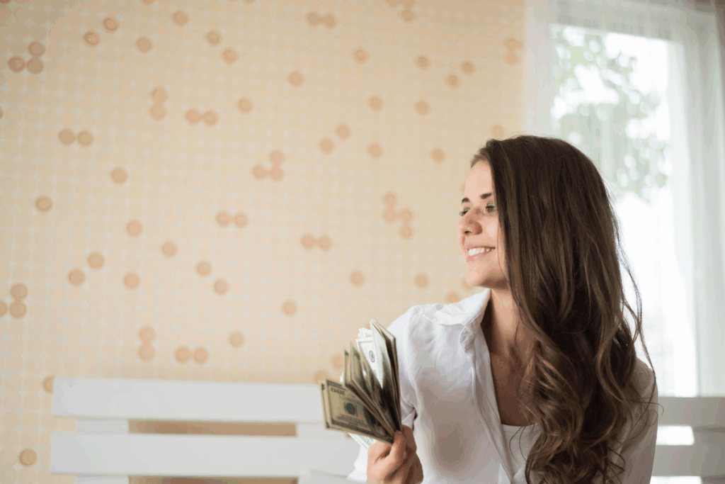 Mulher branca, jovem e sorrindo, segurando uma nota de dólar (dinheiro) enquanto relaxa na cama. A imagem sugere a tranquilidade e a segurança financeira de estratégias de investimento em ano eleitoral.