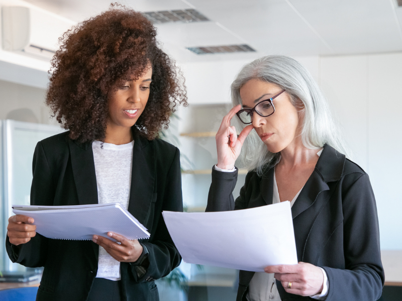 A imagem mostra duas mulheres em um ambiente corporativo, analisando documentos como se estivessem discutindo informações importantes ou revisando relatórios.