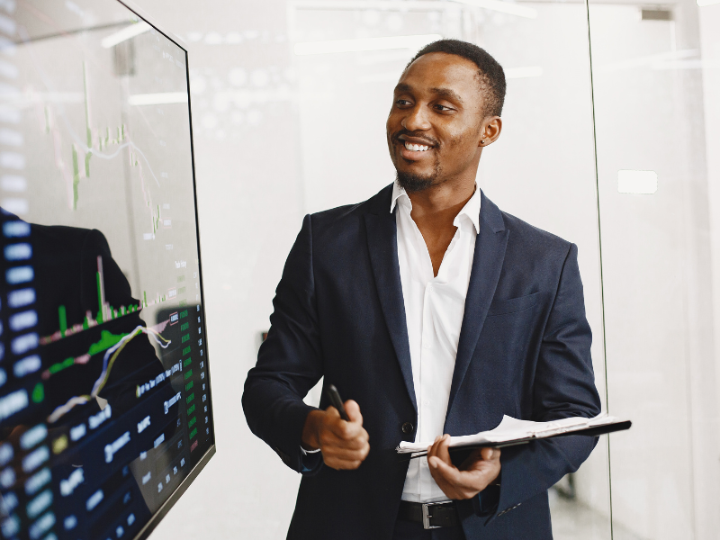 A imagem mostra um homem em um ambiente corporativo, apresentando dados financeiros exibidos em uma tela grande.