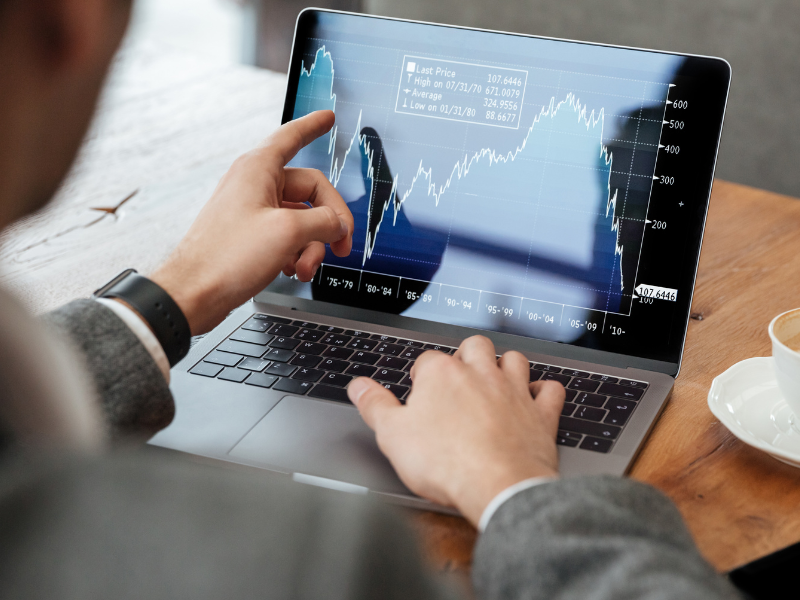 A imagem mostra uma pessoa de costas, vestindo um blazer ou paletó cinza, concentrada em um laptop prateado que exibe um gráfico de mercado financeiro.