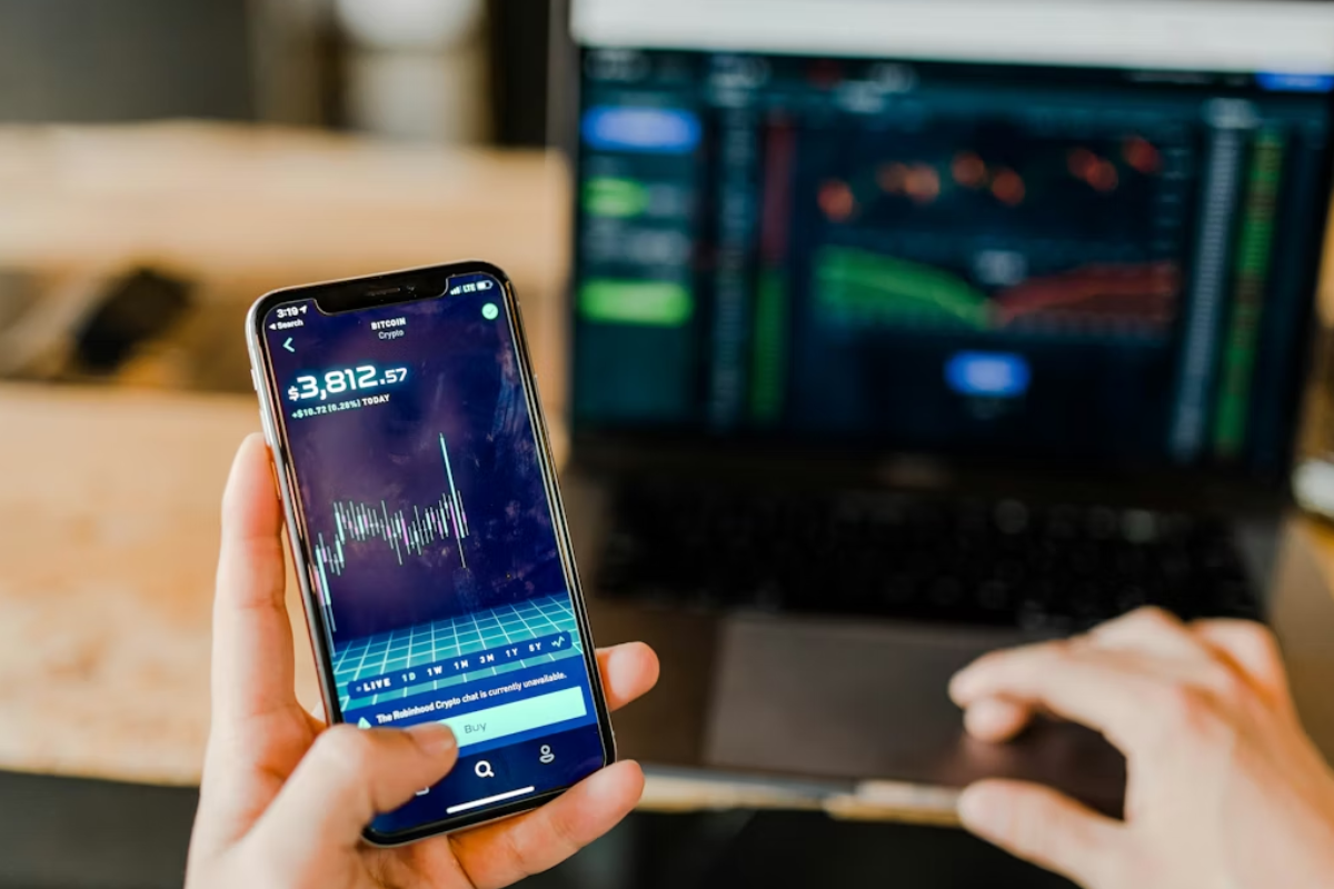 Pessoa sentada em uma escada moderna de madeira e vidro, trabalhando em um laptop prateado, com um telefone celular ao lado, em um ambiente bem iluminado. 